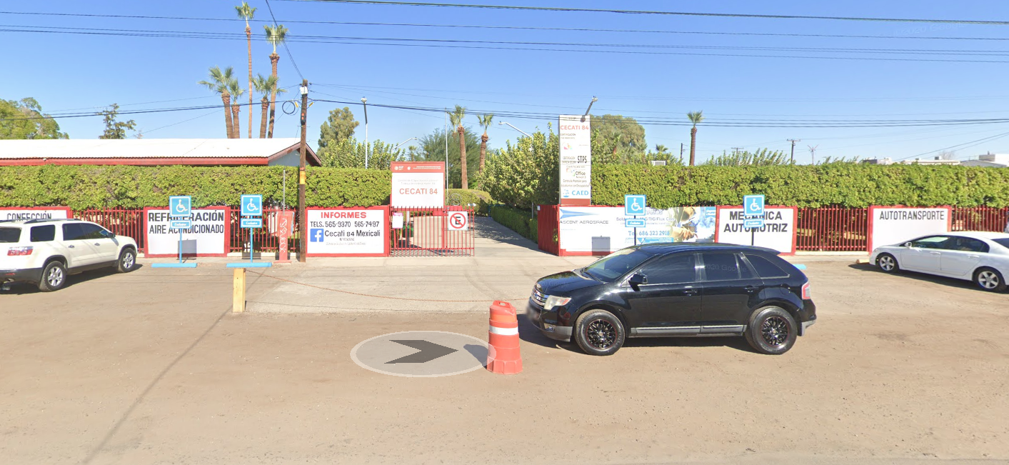 En la fotografía se muestra la fachada del plantel conformada por muros blancos intercalados con rejas rojas. Entre estas paredes y rejas se encuentra el acceso con un cartel donde dice en letras grandes CECATI 84.